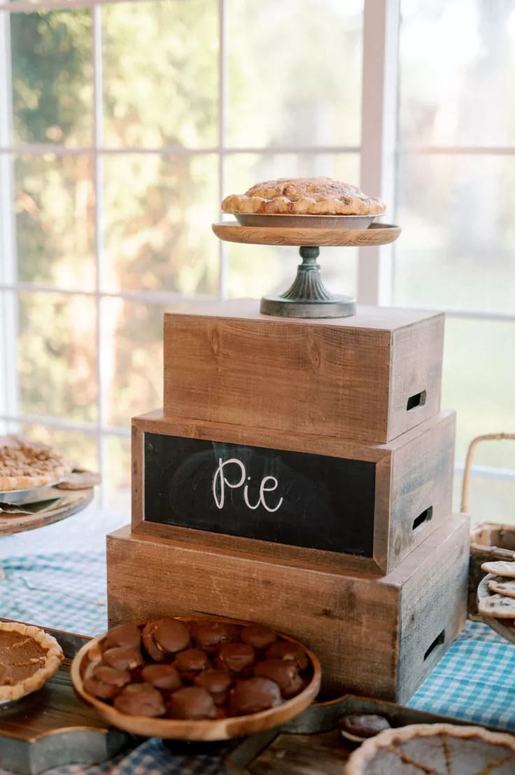 Rustic wedding pie display with stacked wooden crates, a chalkboard sign labeled “Pie,” and various homemade pies arranged on a gingham-covered table.