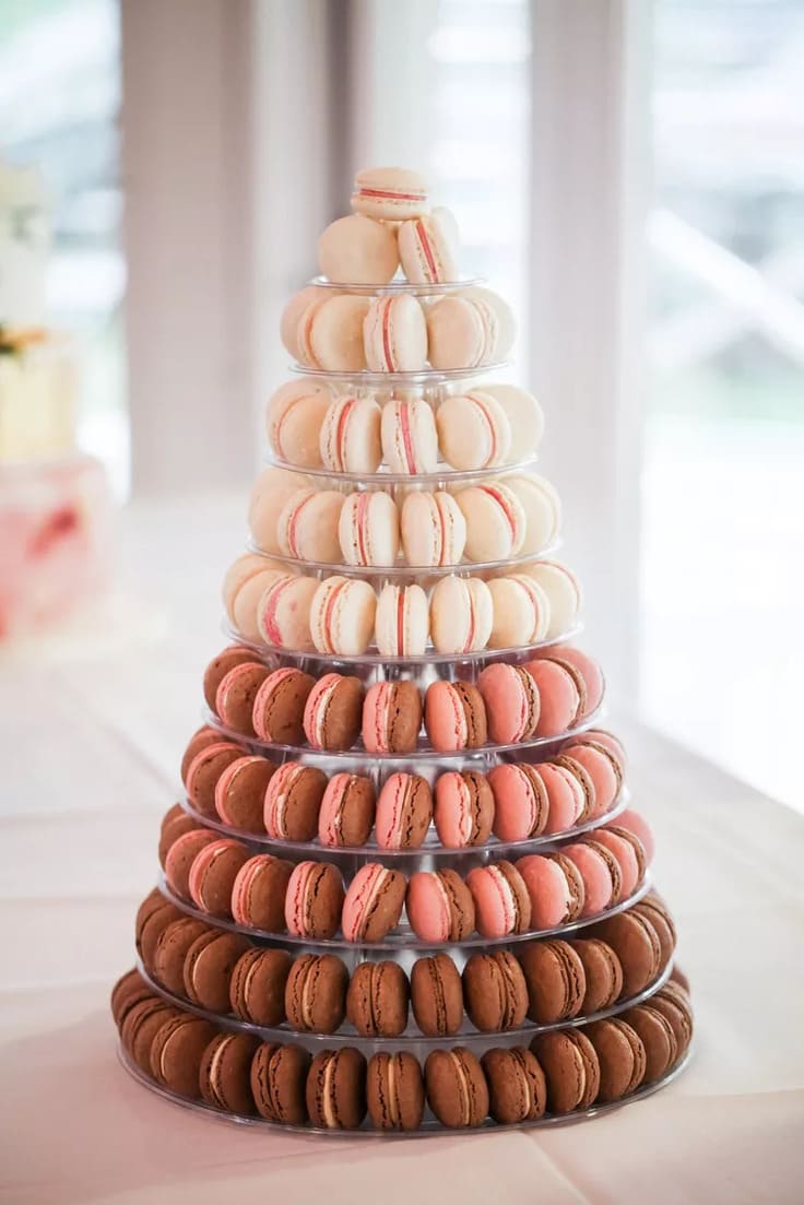 A tower of macarons in shades of chocolate, blush pink, and ivory is arranged on a clear acrylic stand and displayed on a white tablecloth with soft natural light in the background.