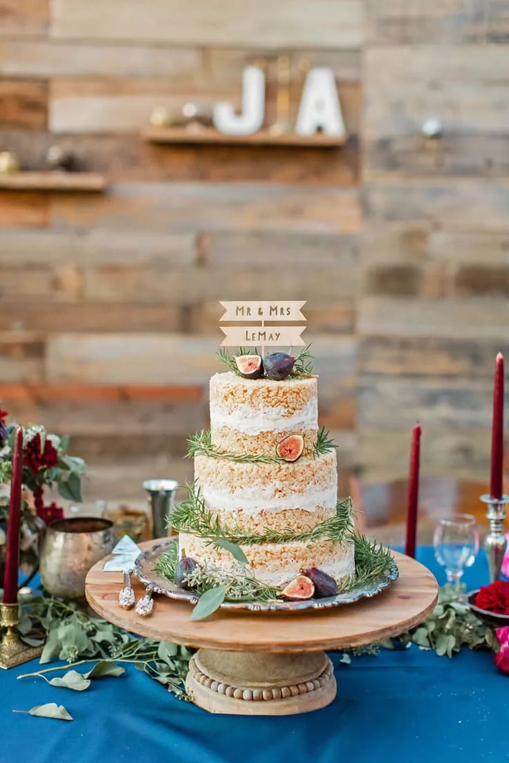 Three-tiered Rice Krispies cake layered with white icing, decorated with figs and sprigs of rosemary, displayed on a wooden pedestal against a rustic wood-paneled backdrop.