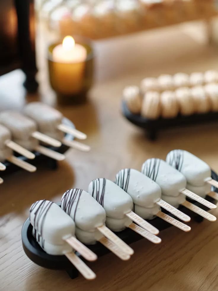 Elegant display of white chocolate-covered popsicles drizzled with dark chocolate, arranged on black trays with personalized wooden sticks, set beside a glowing candle.