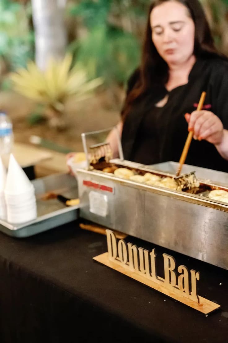 Live doughnut station with a fresh fryer, paper cones, and a wooden “Donut Bar” sign on a black tablecloth, with a vendor preparing golden doughnuts.