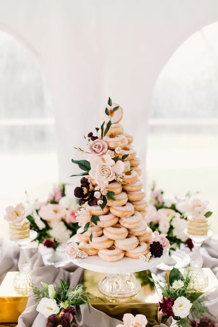 Tower of glazed doughnuts arranged in a cone shape and adorned with soft blush and burgundy flowers, displayed on a glass pedestal with floral arrangements and draped fabric.