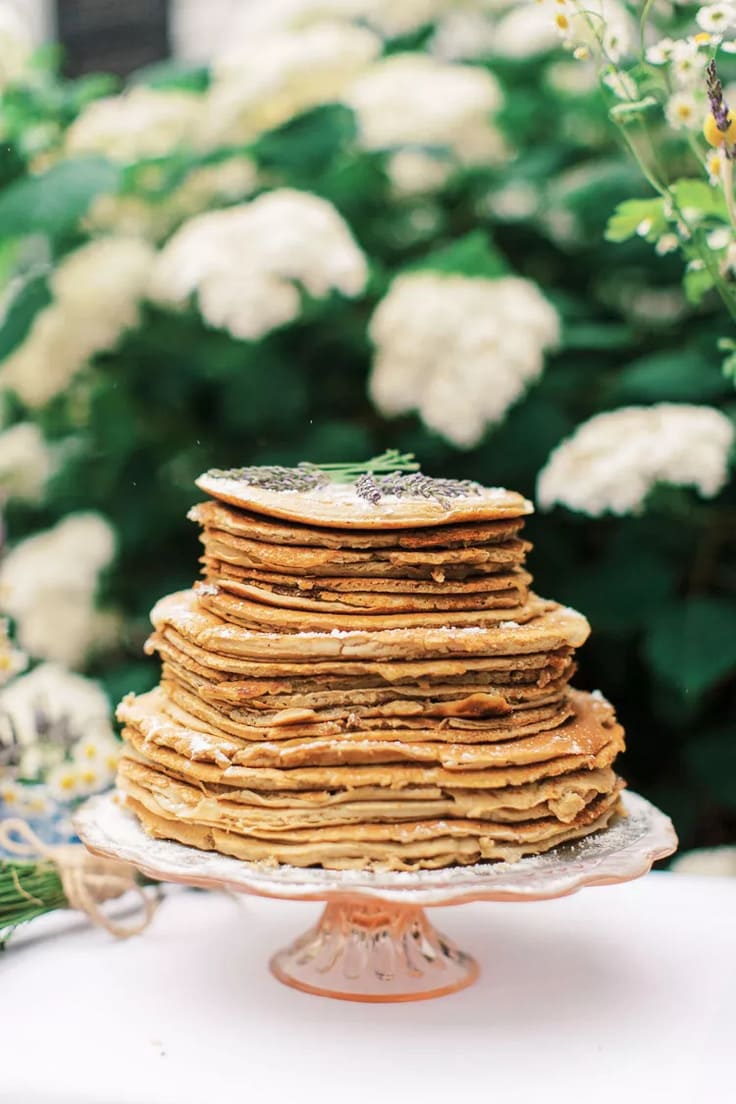A layered stack of golden brown pancakes dusted with powdered sugar and topped with a sprig of lavender is set on a vintage pink glass cake stand with blooming white hydrangeas in the background.