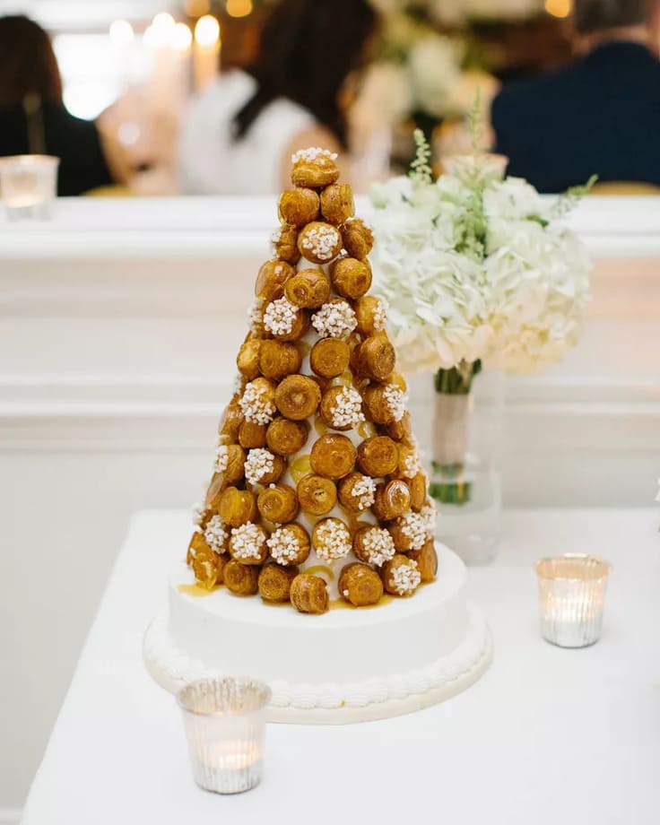 Tall croquembouche tower made of golden cream puffs decorated with clusters of white sugar pearls, set on a round cake base with votive candles and white hydrangeas nearby.