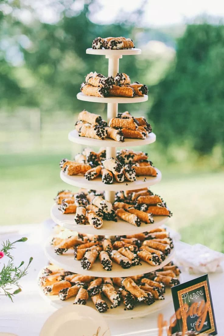 Tower of classic cannoli arranged on a tiered white stand, each filled with creamy ricotta and dipped in mini chocolate chips, set on an outdoor table with greenery in the background.