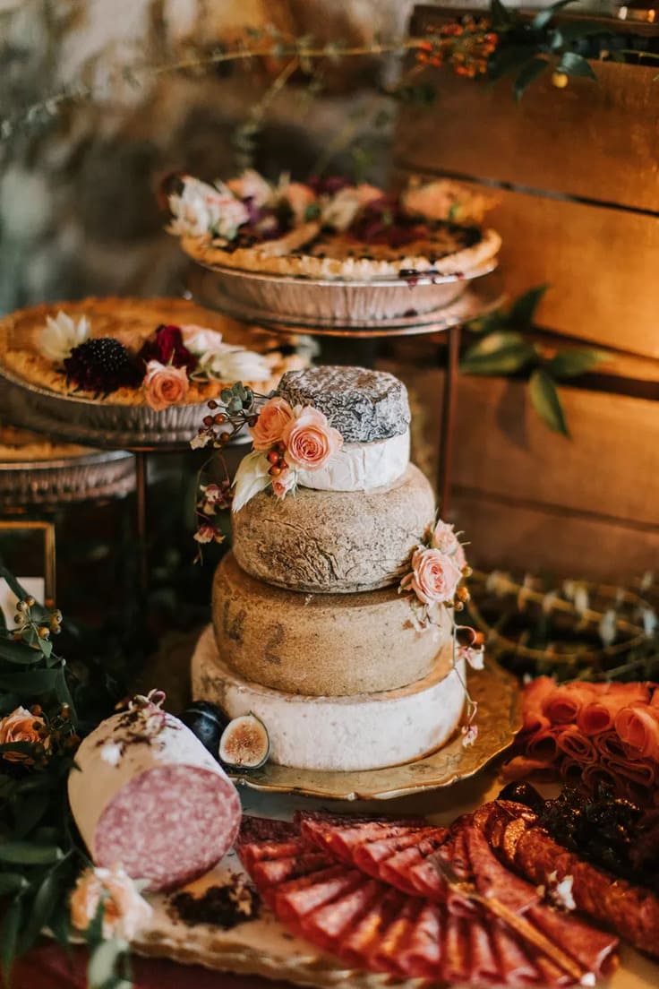 Stacked wheels of artisan cheese styled like a tiered cake, decorated with fresh figs, berries, and blush-toned flowers, surrounded by charcuterie and savory pies.